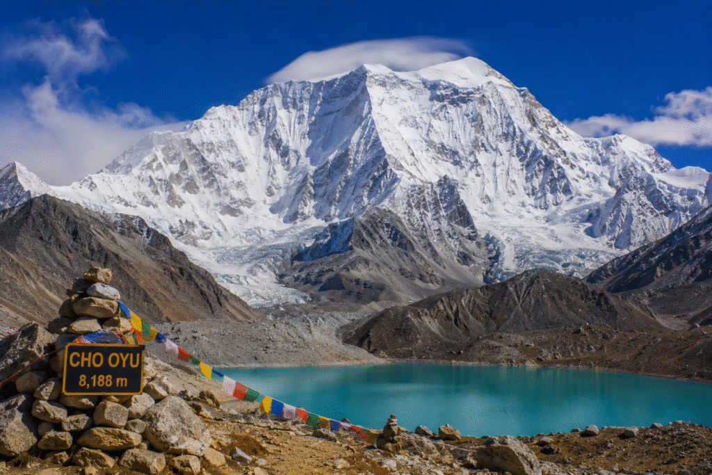 Cho Oyu, the sixth-highest mountain in the world, overlooking a turquoise alpine lake and rugged Himalayan terrain under a bright blue sky