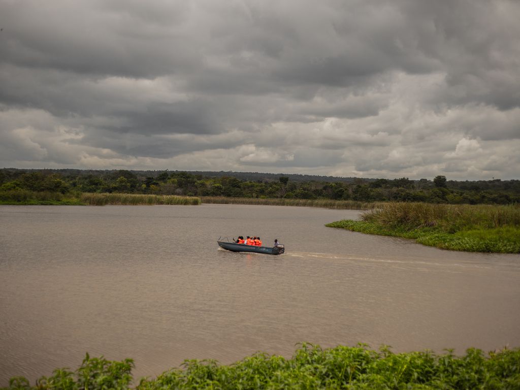 A small boat with people wearing life vests crossing a lake surrounded by lush greenery under a cloudy sky.