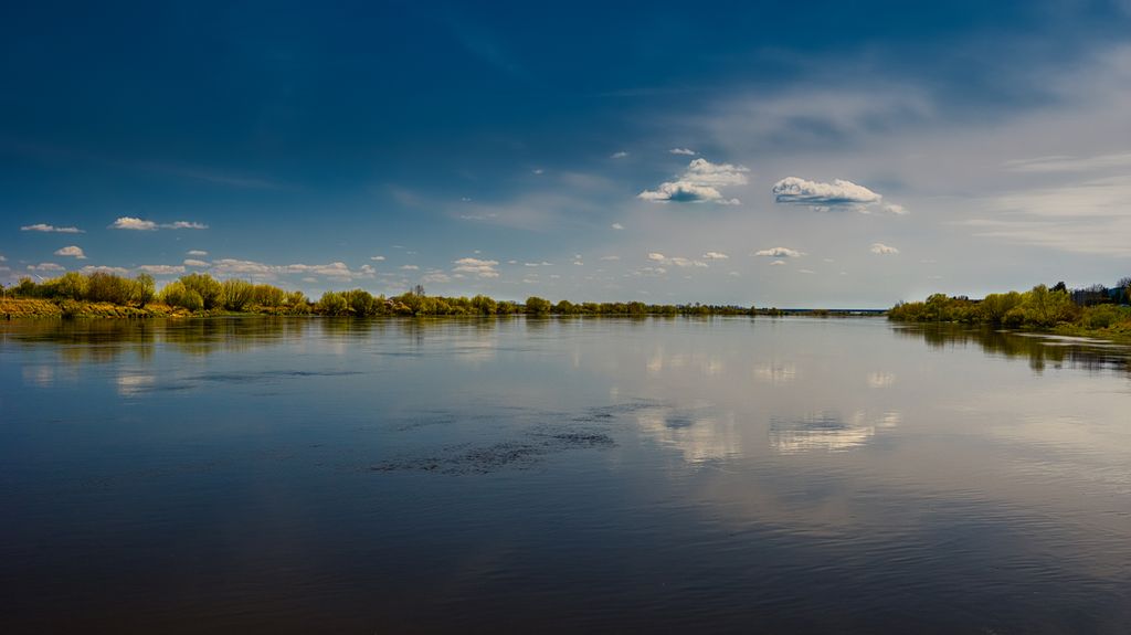 Calm river with green trees reflecting a blue sky and scattered clouds.