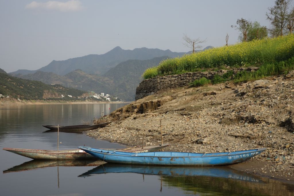 Tranquil riverbank scene with small boats, mountains, and bright green foliage under a clear sky.